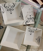 Four white square plates with black and grey ornamental design stacked and arranged on a glass table.