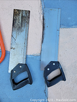 Photo shows two vintage hand saws with black plastic handles, one with a toothed blade showing wear and patina, the other with a smooth blue-coated blade, both for cutting.