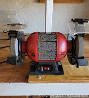 Front view of red bench grinder with dual wheels and clear protective shields on each side, on/off switch visible at the base.