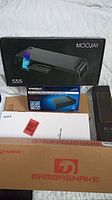 Boxes of items in the lot showing Moojay monitor stand S55 and Sabrent hard drive docking station on top of cardboard boxes.