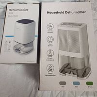 Photo showing two boxed dehumidifiers: a compact white DH-CS01 with dark top and transparent base, and a larger household white dehumidifier with front air vent and 2L water tank capacity.