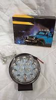 Single LED work light with clear plastic lens, black casing, and screws mounting hardware next to its box with a jeep image on it.
