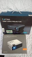 Two boxed digital projectors: TOPTRO Model X5 1080P and Apeman LED projector on a quilted white background.