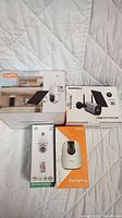 Lot of four unopened smart security cameras in original boxes on white quilted surface.