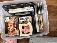 Top view of plastic storage bin containing assorted African American DVDs and CDs, mostly sealed, including a visible DVD with a woman on the cover and various colorful CD spines.