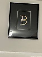 Frontal photo of the framed letter B artwork by Erté hanging on a wall, showing the black frame, dark mat, and detailed artistic stylization of the letter B with integrated figure.