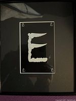 Framed Erte letter E artwork with black matte, showing stylized white letter 'E' composed of figure shapes and wings, with cursive 'e' in corners and artist signature.