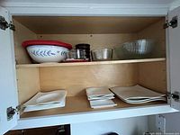 Upper shelf of cabinet showing ceramic bowls with red lids, stacks of glass storage bowls with lids, large clear glass serving bowl.