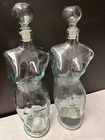 Front view of two clear glass female torso shaped bottles with round stoppers designed for oil or vinegar.