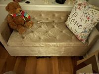 Overall front view of beige tufted storage bench with 'Love' pillow and teddy bear on top.