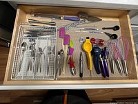 Photo of an open drawer with stainless steel silverware including forks, spoons, butter knives, a grater, tongs, citrus juicers, whisks, and silicone spatulas organized neatly.