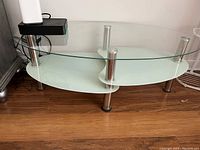 View of the entire three tiered table, showing the clear glass top with curved frosted glass shelves below, supported on chrome metal legs.