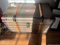 Top view of closed trunk showing crocodile pattern exterior, leather-like straps with rivet accents, black metal corner protectors, and central locking latch. 'Fragile' sticker is present on lid.