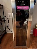 Closed wine fridge showing stainless steel frame and glass door with a long vertical metal handle and a power cord plugged in.