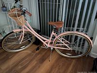 Left side view of the pink bicycle showing the step-through frame, front wicker basket, handlebar with bell and brake cables, brown saddle, rear rack, and front and rear fenders.