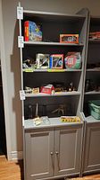 Front view of gray wooden bookcase with three shelves holding model kits and toys, two-door cabinet below