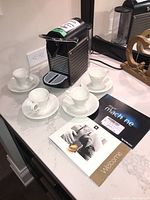Nespresso machine with 4 white espresso cups and saucers with manuals on white countertop.