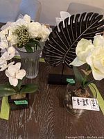 Photo showing three main faux plants/flowers in glass vases and decorative leaf sculpture on dark wood table.