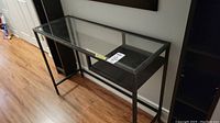 Front view of rectangular hall table showing clear glass top on black metal frame and storage cubby below.