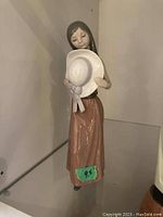 Front view of the Lladro figurine holding a white hat, showing the glossy brown dress and long brown hair.