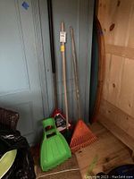 Photo showing three rakes standing against a blue door and include a new pack of green leaf bags with a rigid frame.