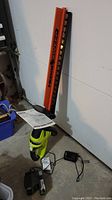 Full view of Ryobi electric hedge trimmer with orange blade guard, lime green and black body, batteries, charger and manual on floor.