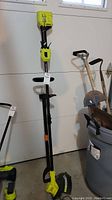 Full view of Ryobi cordless string trimmer leaning against wall with Fiskars weed puller in background