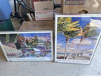 Two framed Art Fest by the Sea posters from 1999 and 2000, showing coastal scenes with boating and palm tree imagery. Posters positioned side by side on a floor among storage boxes.