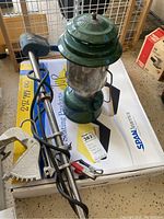 Photo showing green vintage lantern, small boat motor with cables, and white metal boat seat base on a box.