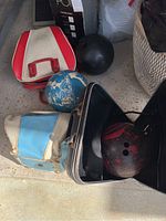 Photo showing three bowling balls with zippered cases on a floor. Bowling balls include a black and red one, blue and white marbled one, and a solid black one. Cases are red/white, light blue/white, and black in color and appear weathered.