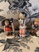 Photo showing two ceramic children's statues: Asian girl kneeling holding fan, and a young girl in orange dress by a lantern post, seated on a floral cover surface.