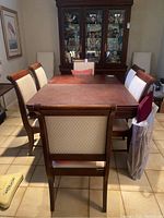 Six wooden dining chairs around a dining table showcasing cream colored diamond-patterned cushioned seats and backrests. Chairs show moderate wear, some stains clear on cushions.