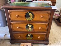 Front view of a wooden side table with three drawers. Each drawer front is painted with a circular illustration of a golfer in action. Wooden knobs on each drawer. Evidence of scratches and wear on the surface.