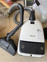 Top view of white Miele Turbo Plus canister vacuum with attached hose and black floor head on speckled floor surface.