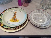 Photo showing a stack of assorted decorative platters and plates, including a yellow floral rim platter with a central illustration and a black and white etched design plate.