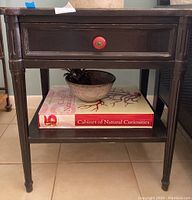 Front view of dark wooden side table showing single drawer with red knob and lower shelf holding books and bowl (contents not included).
