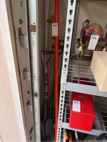 Yard tools stored upright between shelving and wall, visible long orange broom handle and wooden handle shovel with metal scoop and Kodiak label.