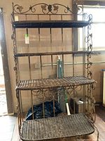 Full view of rattan and metal bakers rack showing three rattan shelves and decorative metal grapevine design on frame.