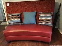 Front view showing the red curved bench with three pillows placed on the seat, highlighting the faux leather seat and fabric backrest.