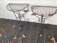 Two matching semi-circular metal tables with grid tops and decorative metal leaves on three-legged bases, placed on a porch floor with scattered dry leaves.