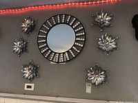Six silver tone metal wall flowers mounted on a gray wall around a round mirror. The flowers all have a layered petal design with rhinestones on the petals.