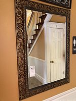 Front view of rectangular beveled wall mirror with ornate carved frame hanging on wall, showing mirror reflection of staircase and door.
