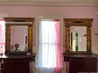 Two matching antique-style wall mirrors placed side by side on top of wooden cabinets in a room with pink curtains and light walls. Both mirrors have ornate finish and visible damage.