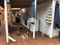 Photo shows four patio umbrellas standing closed on a wooden deck, including a blue umbrella with decorative border, a striped umbrella, and a tiki-style umbrella.