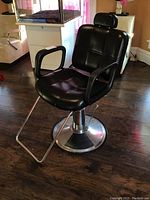 Black professional salon chair with padded seat, armrests, adjustable headrest, chrome round hydraulic base, and footrest on dark wood floor.