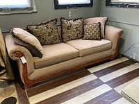 Full view of beige upholstered sofa with carved wood frame, decorative cushions on top, positioned against grey wall with windows and patterned floor beneath.