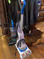 Two upright vacuum and carpet cleaner units standing on wooden floor. Hoover Power Dash Pet is white and blue with transparent water tank; Bissell PowerForce vacuum is black and blue with clear dirt container.