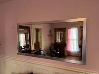 Rectangular silver toned framed wall mirror mounted on a lilac wall with wainscoting. Reflection shows salon setting with chairs and windows.