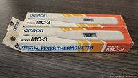 Two Omron MC-3 digital fever thermometers in their original packaging on a dark surface, each showing 98.6°F on their digital screens.