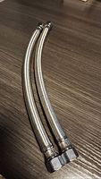 Two new stainless steel braided water supply hoses with hexagonal nuts on both ends placed on a wooden surface.